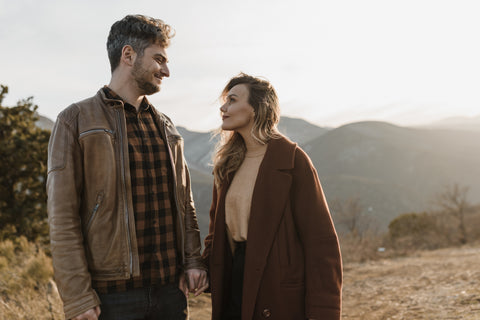 A white man with salt and pepper hair in his late thirties dressed in a brown leather jacket with a brown and grey check shirt looking over to a pretty blonde white woman in a rust coloured wool coat and soft brown sweater. They are stood in front of a vista of mountainous terrain with shrubby and heath like plants. 