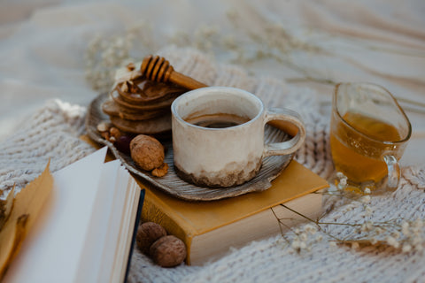 A rustic still life set up of a cup of tea in a rustic pottery cup and saucer placed on an old hardback book. Surrounding it are dried flowers, nuts, a honey drizzler and glass jug of honey. These are all placed on top of a white knitted blanket 
