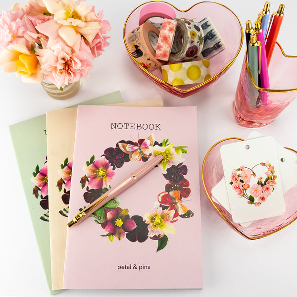 notebooks featuring a flower garland design on a desk with heart shaped glass bowls filled with MT was tape, Ballograf pens in a rainbow of colours, gift tags with a heart designed in rose buds and a vase of garden roses.