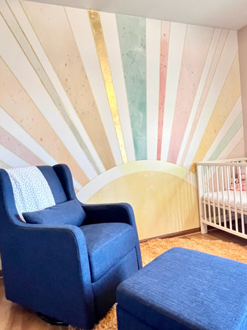 Children's nursery with blue chair, crib and rainbow color sun themed wallpaper mural.