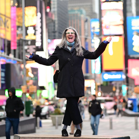 Natalia, dressed in a dark gray car coat, black pants and shoes, and black and white striped socks, is standing on a platform in Times Square with her arms outspread and a huge smile (or fear?) on her face.