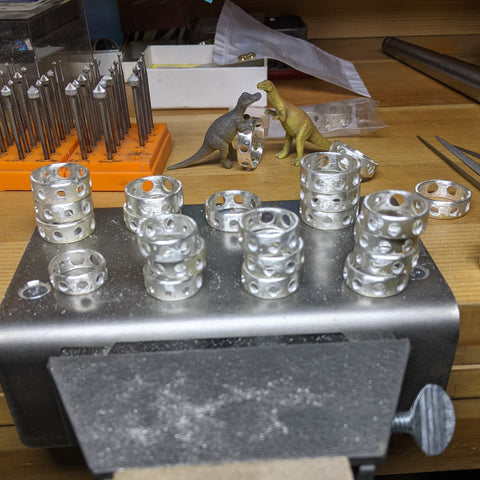 Stacks of knitting needle gauge rings on Natalia's work bench, with two of her little dinosaurs in the background moving rings.