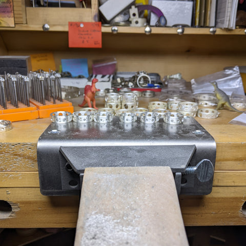 Rows of unfinished silver knitting needle gauge rings are sitting on a wood jeweler's bench, with a small red plastic dinosaur in the background.