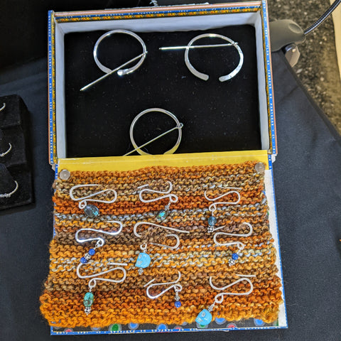 A cigar box open with a black velvet pad inside the box at the top of the picture, and with a knit swatch in shades of orange attached to the lid at the bottom of the image. In the box are 3 penannular shawl pins made in silver, and on the knit swatch are 3 small shawl pins that resemble a squashed "S" and have lapis lazuli and turquoise beads hanging from them.