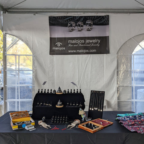 The Malojos Jewelry booth at Wool and Folk, featuring a table with a black tablecloth, upon which there are jewelry displays on risers, and a sign hanging in the back of the white tent with picture of the Knitting needle gauge rings and the Malojos Jewelry logo.