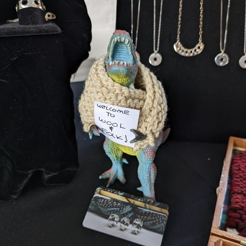 A blue plastic T-Rex wearing a cream colored shawl, and holding a sign that reads: welcome to wool and folk"