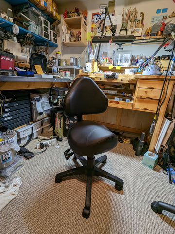 Weird black office chair with a seat shaped like a wide bicycle seat, in the middle of a crowded jewelry studio.