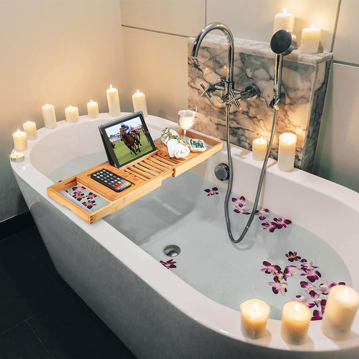 Bath tub filled with water and flower petals with candles around the edge and a bath tray across the center holding a tablet, a phone, and a glass of wine..