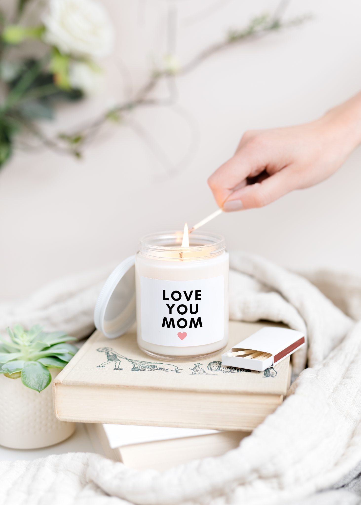 Love You Mom Candle in jar with white lid leaning against its side is being lit on top of a stack of books against a white background.
