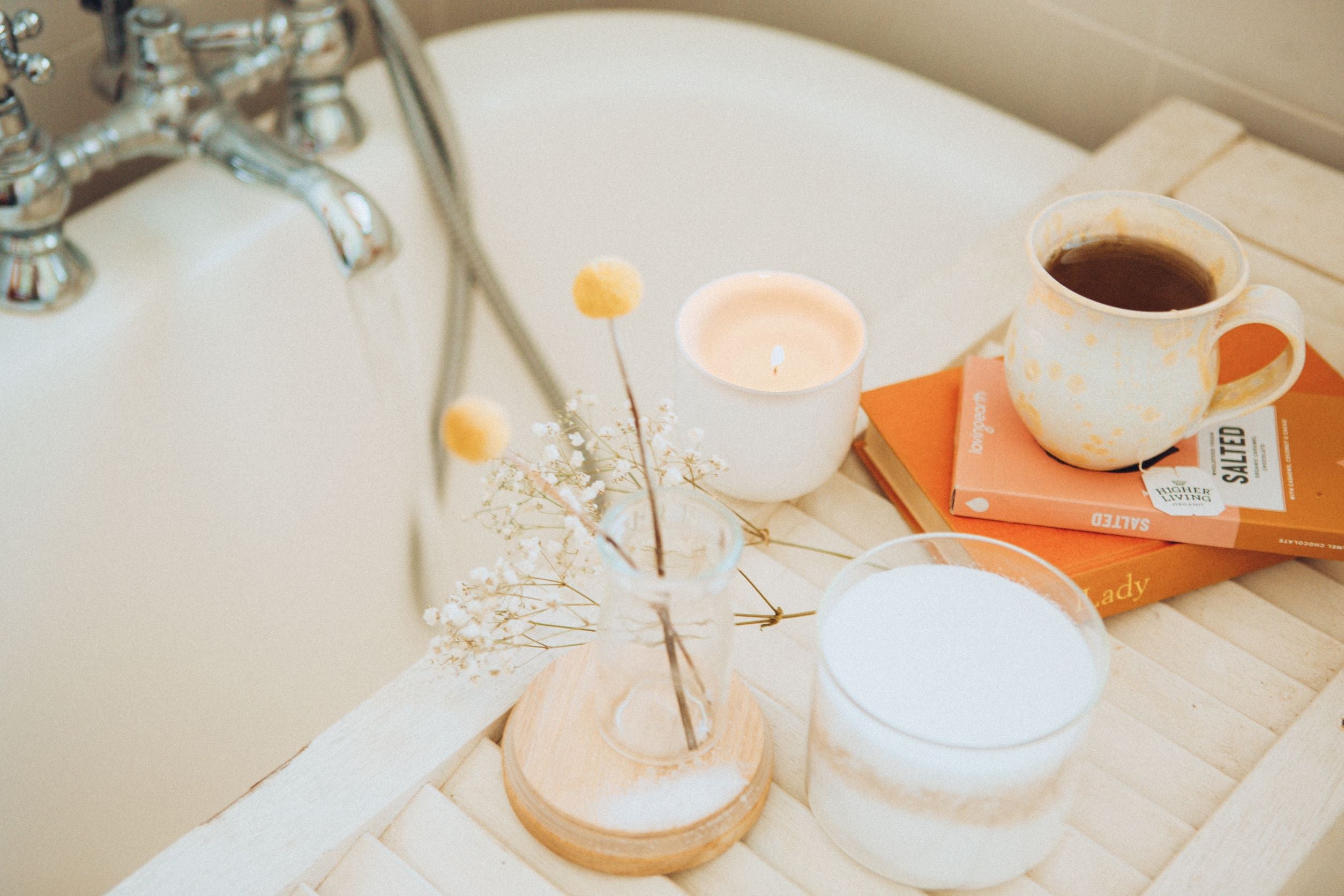 Image of a bath tray over a bath tub with items on it - cup of tea, books, candle, flowers.