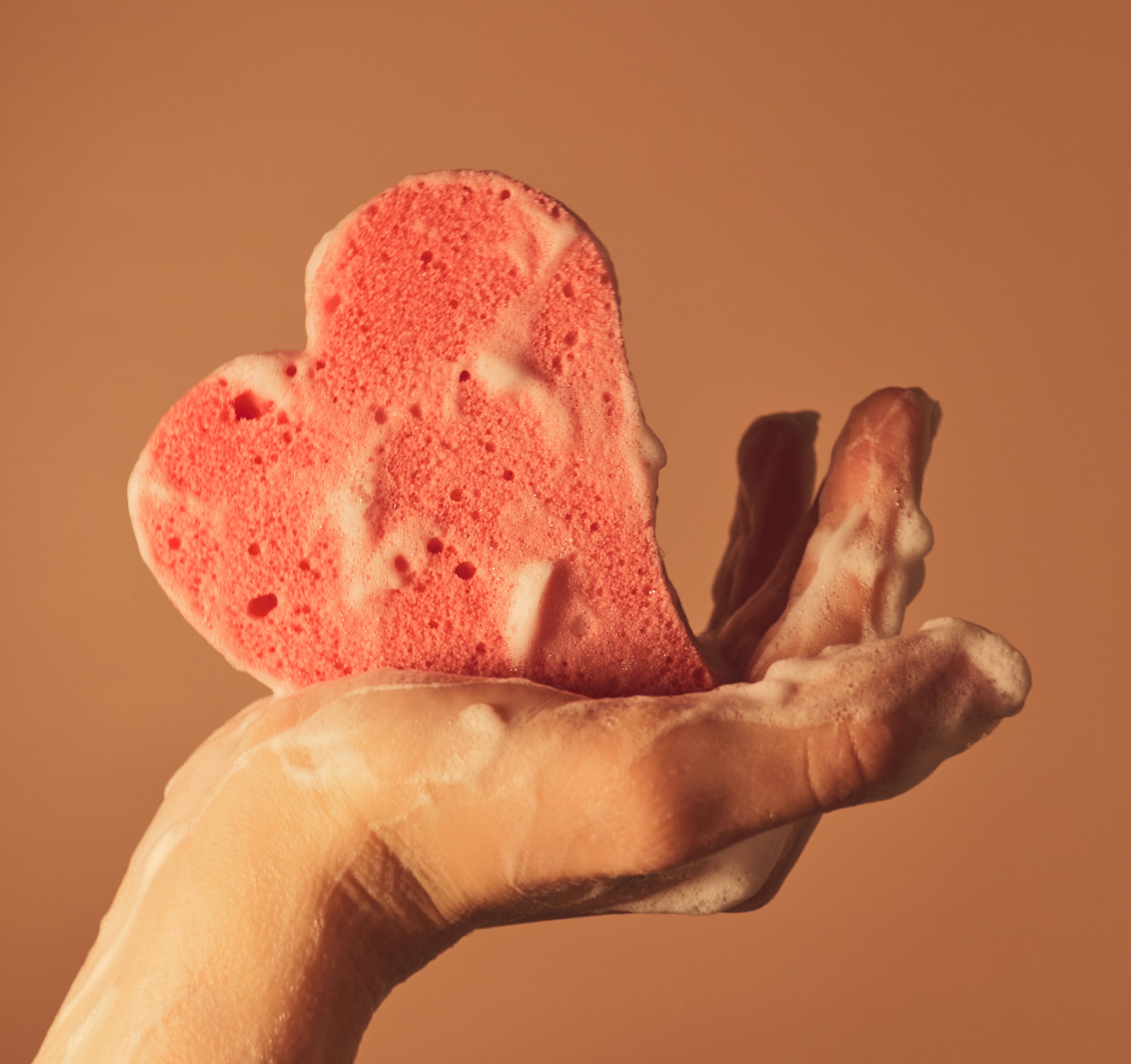 Image of a hand holding a pink sponge that is shaped like a heart with soap suds on it.
