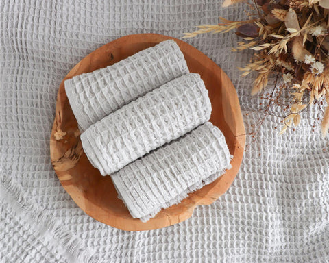 Image of three light grey cotton waffle towels rolled up in a wooden bowl, with a dried flower bouquet just seen in the top right hand corner