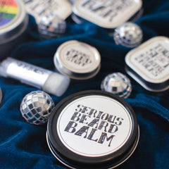 Serious Lip Balm products like hand stuff, lip balm and beard stuff sit on blue velvet with a disco ball.