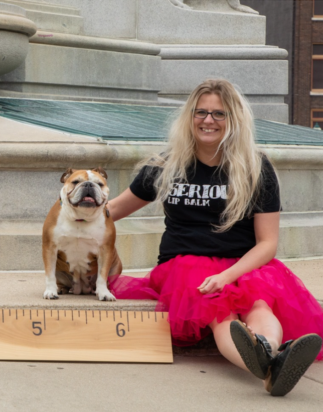 Woman with long blonde hair in a black tshirt with Serious Lip balm on it sits next to a brown and white English bulldog, both are smiling.