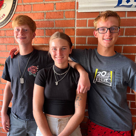 three siblings stand in front of a brick wall, smiling at the camera.  Two boys on each side have their arms around their sister in the middle