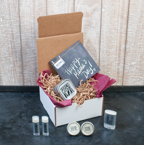 White box with brown crinkle paper and an assortment of all natural and handmade lip care and skin care products on a black surface with wood background