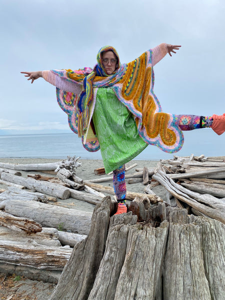 photo of a young woman standing on a large tree stump on a beach. She wears many shawls and scarves and colorful clothes. 
