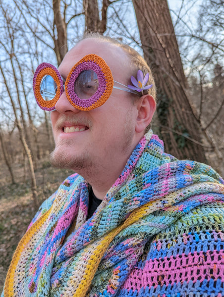 Photo of a young blonde man wearing a colorful crocheted shawl and sunglasses. 