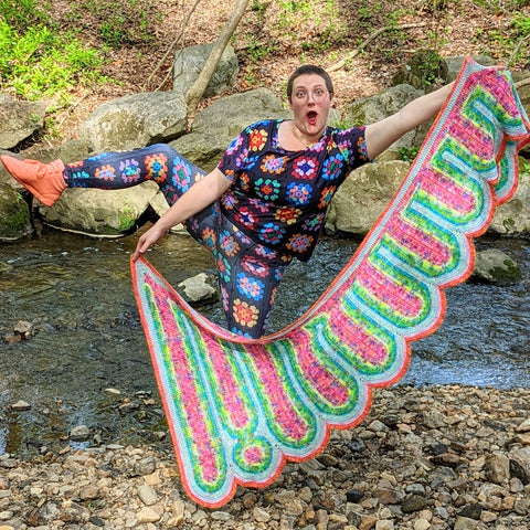 A woman stands on a creek bed, one leg in the air. She holds an extremely long shawl in her arms and is wearing a suit made of granny squares. 