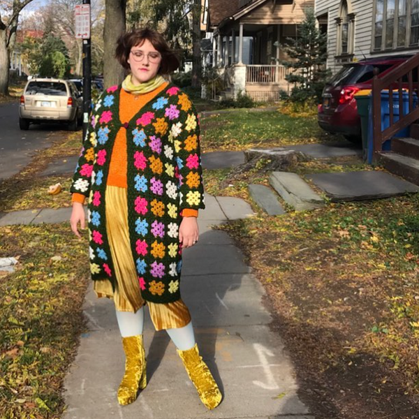 I woman stands on the sidewalk, wearing a brightly colored granny square coat and gold, crushed velvet accessories.