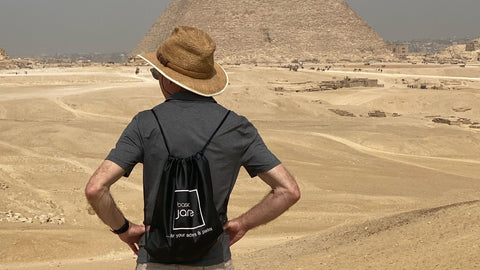 Man standing in front of Egyptian pyramid wearing a Basic Jane CBD brand back pack and hat.