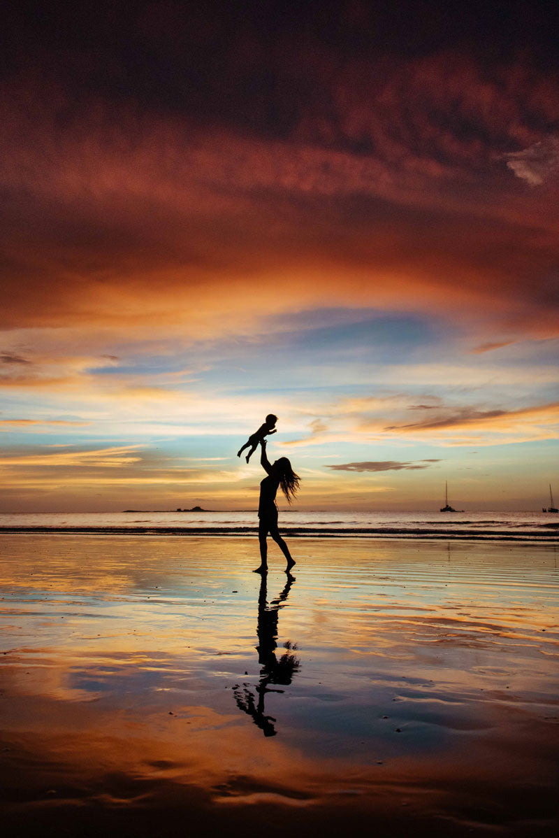 Mom throwing baby into the air during a gorgeous sunset in Costa Rica. Gorgeous pink sunset reflections off the sand in Tamarindo Costa Rica. Photographed by Kristen M. Brown, Samba to the Sea for The Sunset Shop.