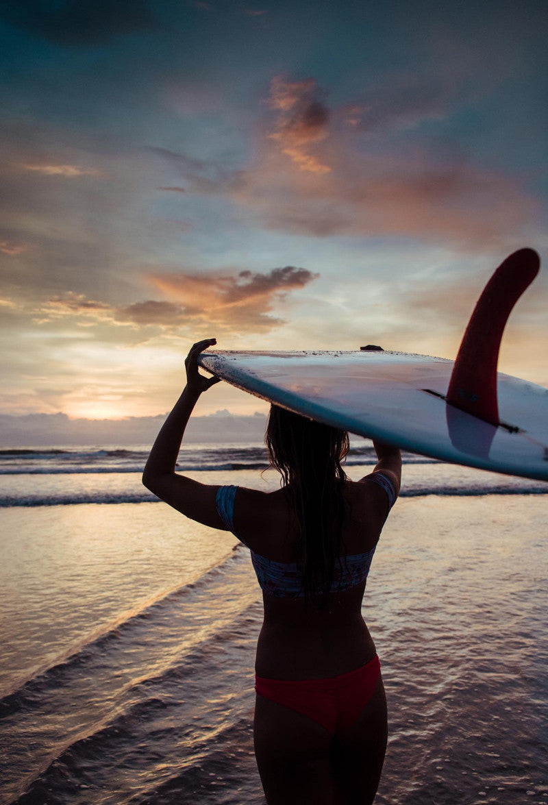 Surfer girl holding a surfboard on her head in Tamarindo Costa Rica during a pretty sunset. Photographed by Kristen M. Brown, Samba to the Sea x The Sunset Shop.