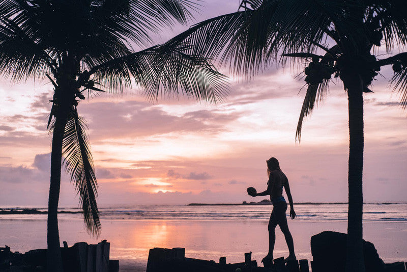 Surfer girl walking between palm trees on the beach in Tamarindo Costa during a pink sunset. Photographed by Kristen M. Brown, Samba to the Sea x The Sunset Shop.