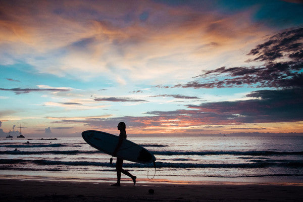 Surfer girl walking on the beach during a pastel sunset in Tamarindo, Costa Rica. Photographed by Kristen M. Brown, Samba to the Sea for The Sunset Shop.