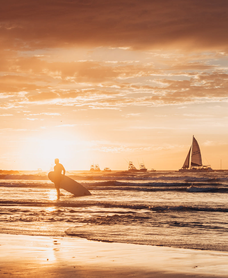 Surfer walking on the beach during sunset in Tamarindo Costa Rica. Photographed by Kristen M. Brown, Samba to the Sea for The Sunset Shop.