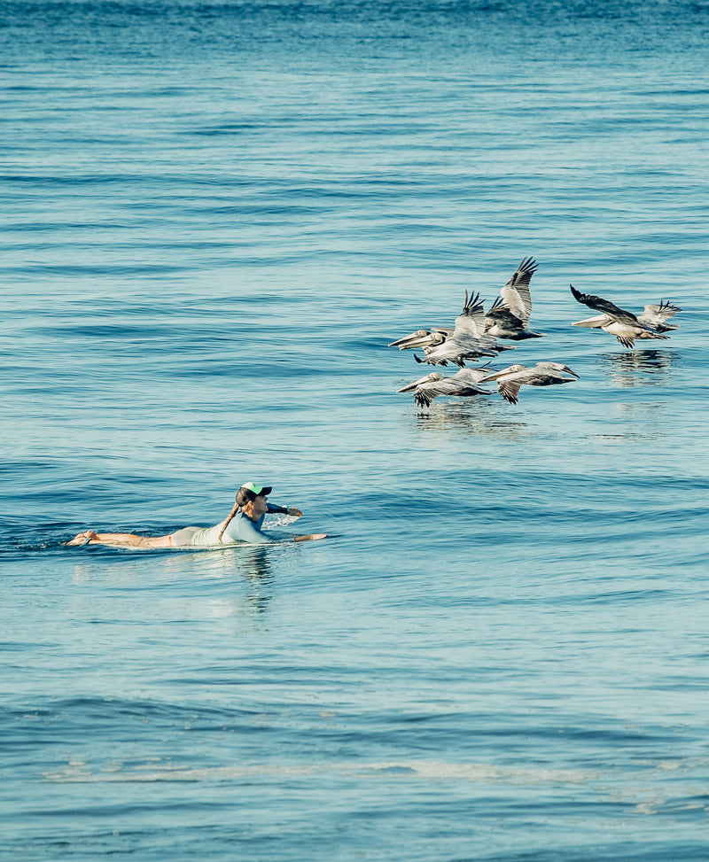 Surfer girl paddling with pelicans in the Pacific Ocean in Costa Rica. Photographed by Kristen M. Brown, Samba to the Sea for The Sunset Shop.