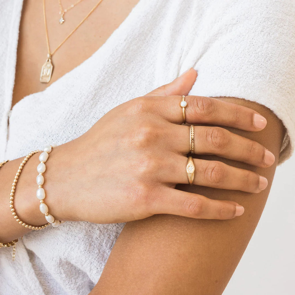 Woman in a white top wearing two necklaces, four gold rings on one hand and two stretch beaded bracelets.