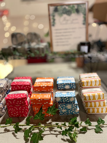 College Green soaps wrapped in brightly coloured Cambridge Imprint paper on an indoor market stall. There's ivy in the foreground and an out of focus sign in the background