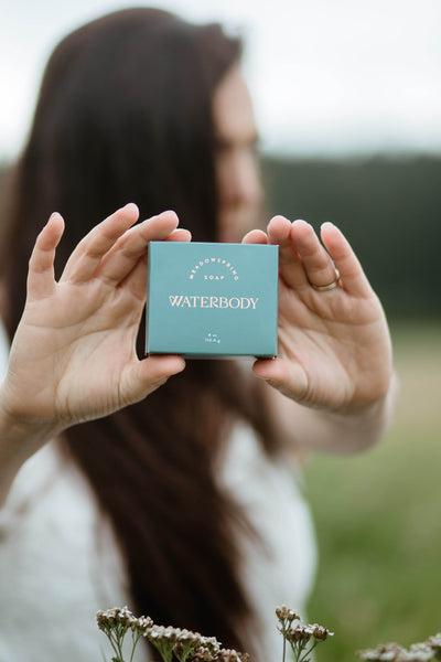 A woman standing in a meadow holds a bar of Meadowspring Soap with two hands