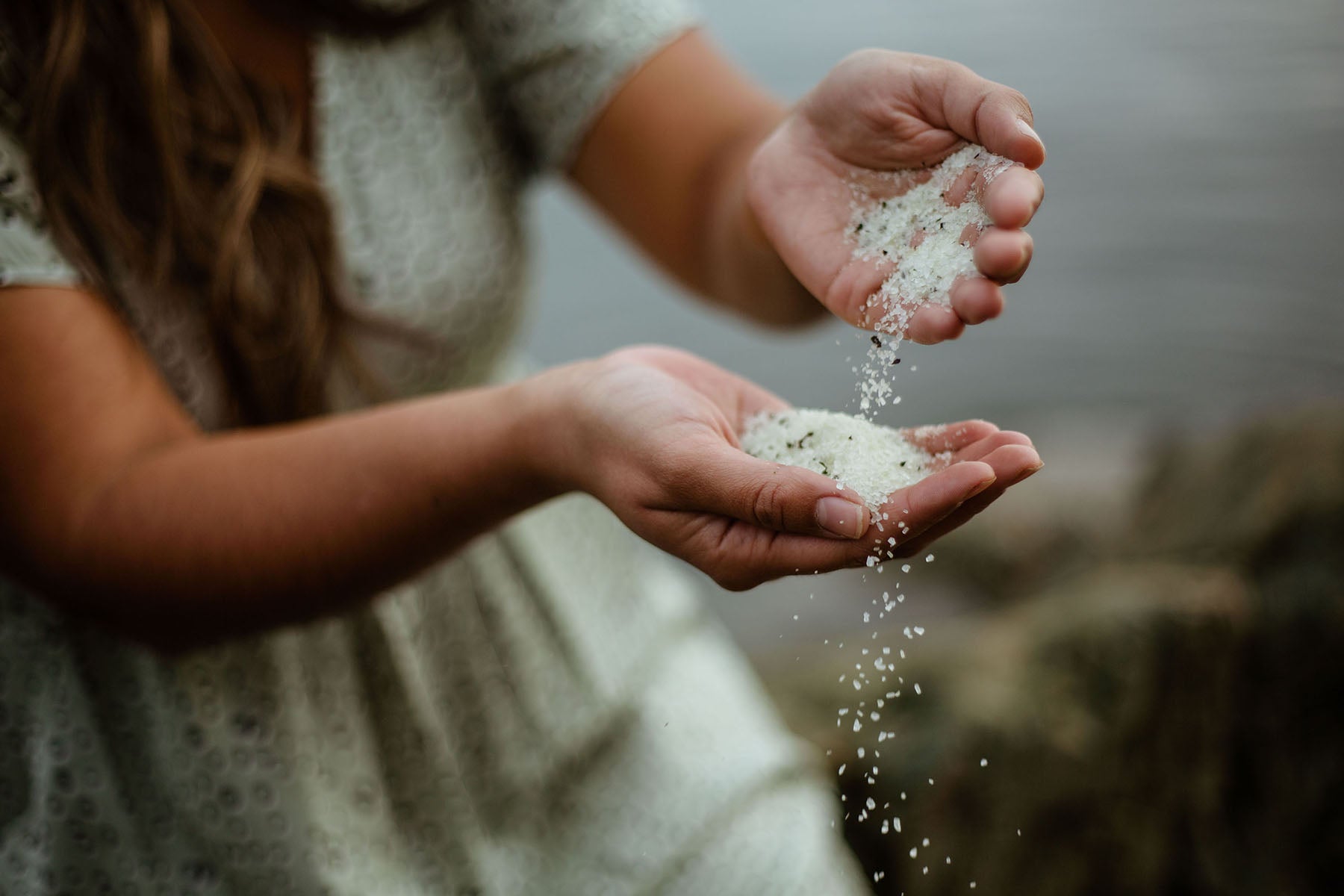 A woman sits beside the ocean and pours a sea salt bath soak from one palm into another
