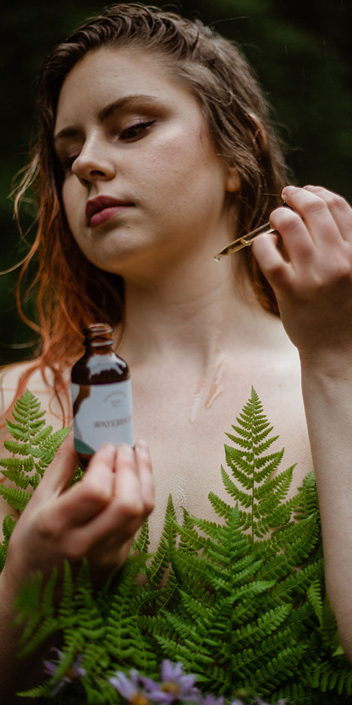 A beautiful woman surrounded by lush ferns applies evergreen body oil to her skin