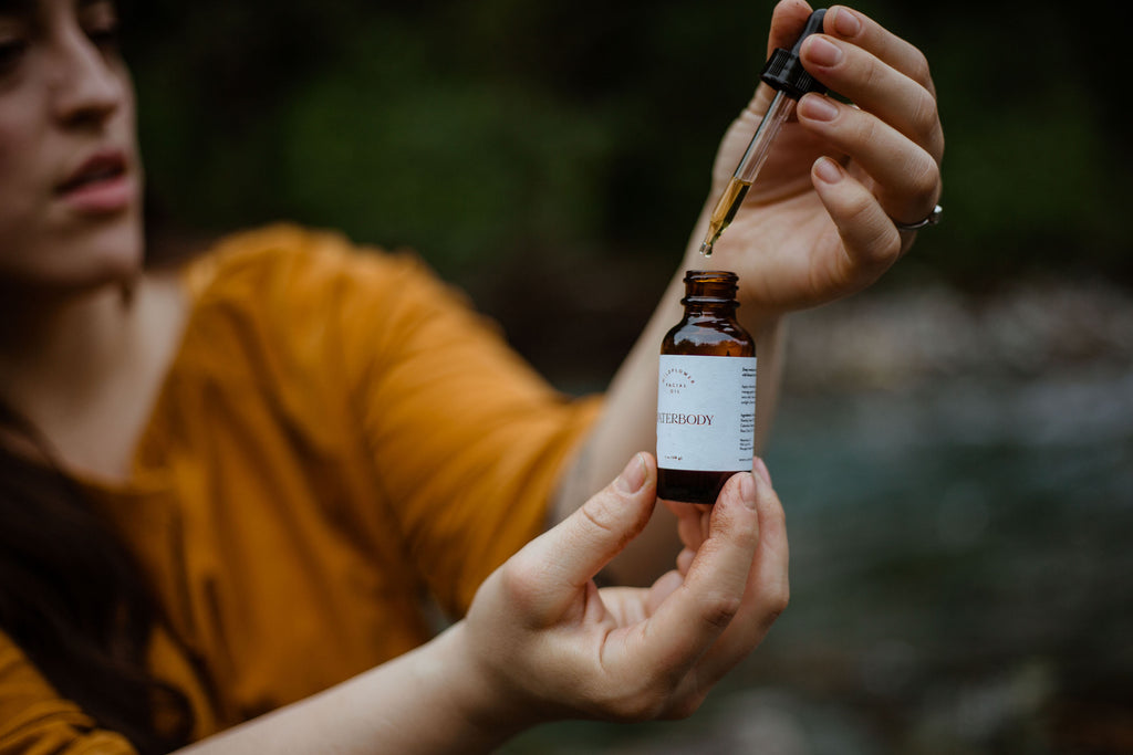 A woman holds a bottle of Waterbody Wildflower Facial Oil and extracts some oil in a dropper near a stream in Alaska