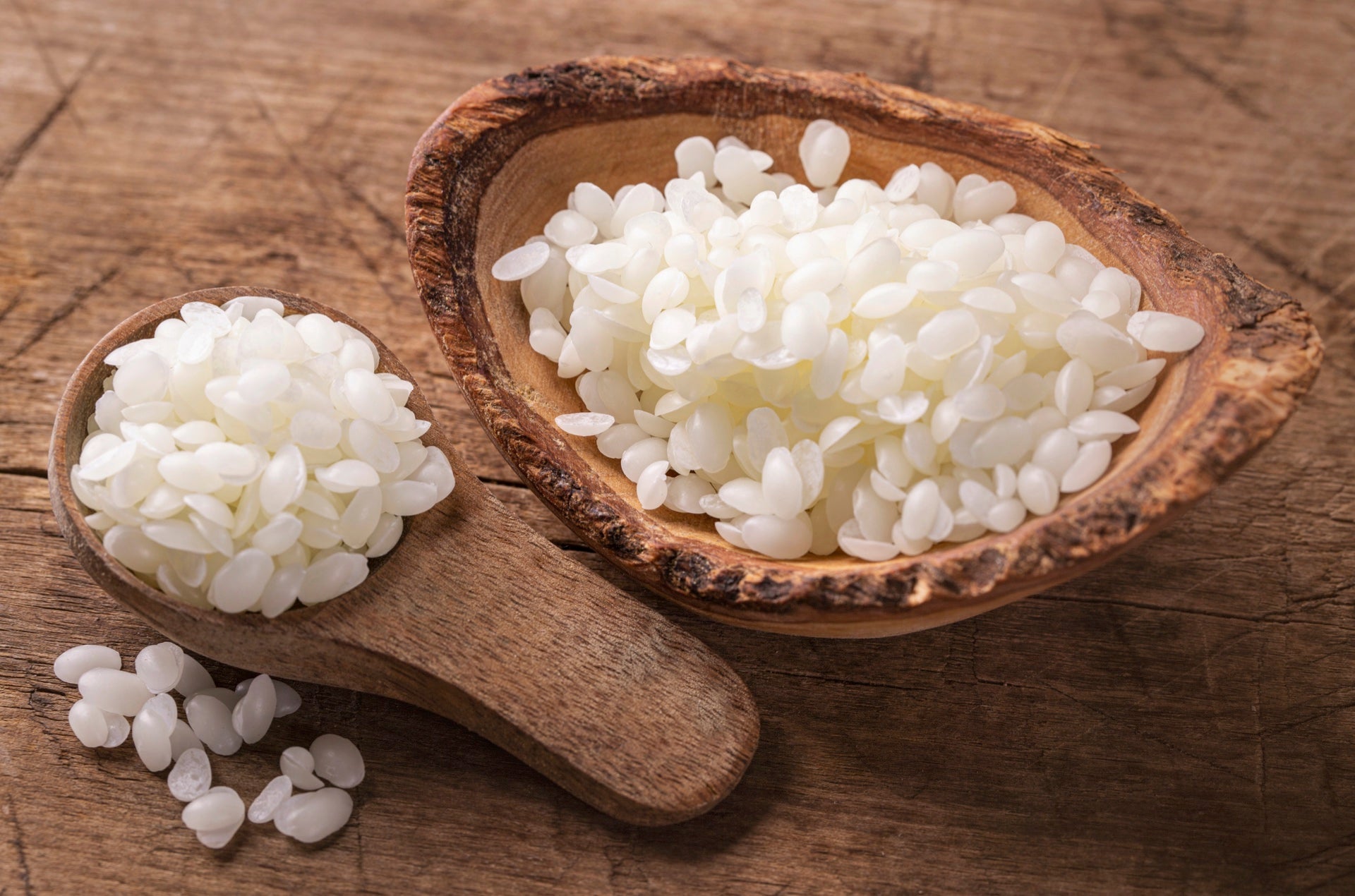 A pile of small white beeswax pellets on a wooden surface, with a few loose pellets scattered around. 