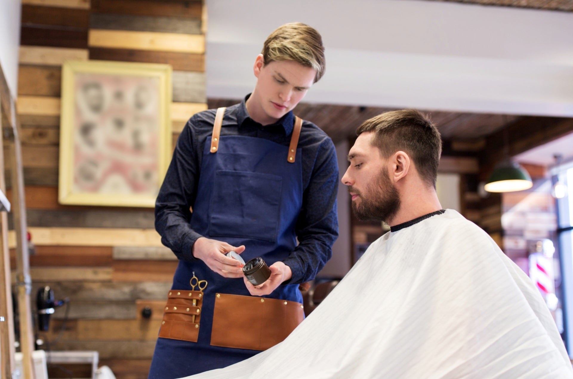 A professional barber in a blue apron holds a jar of hair product, showing it to a male customer seated in the barber chair.