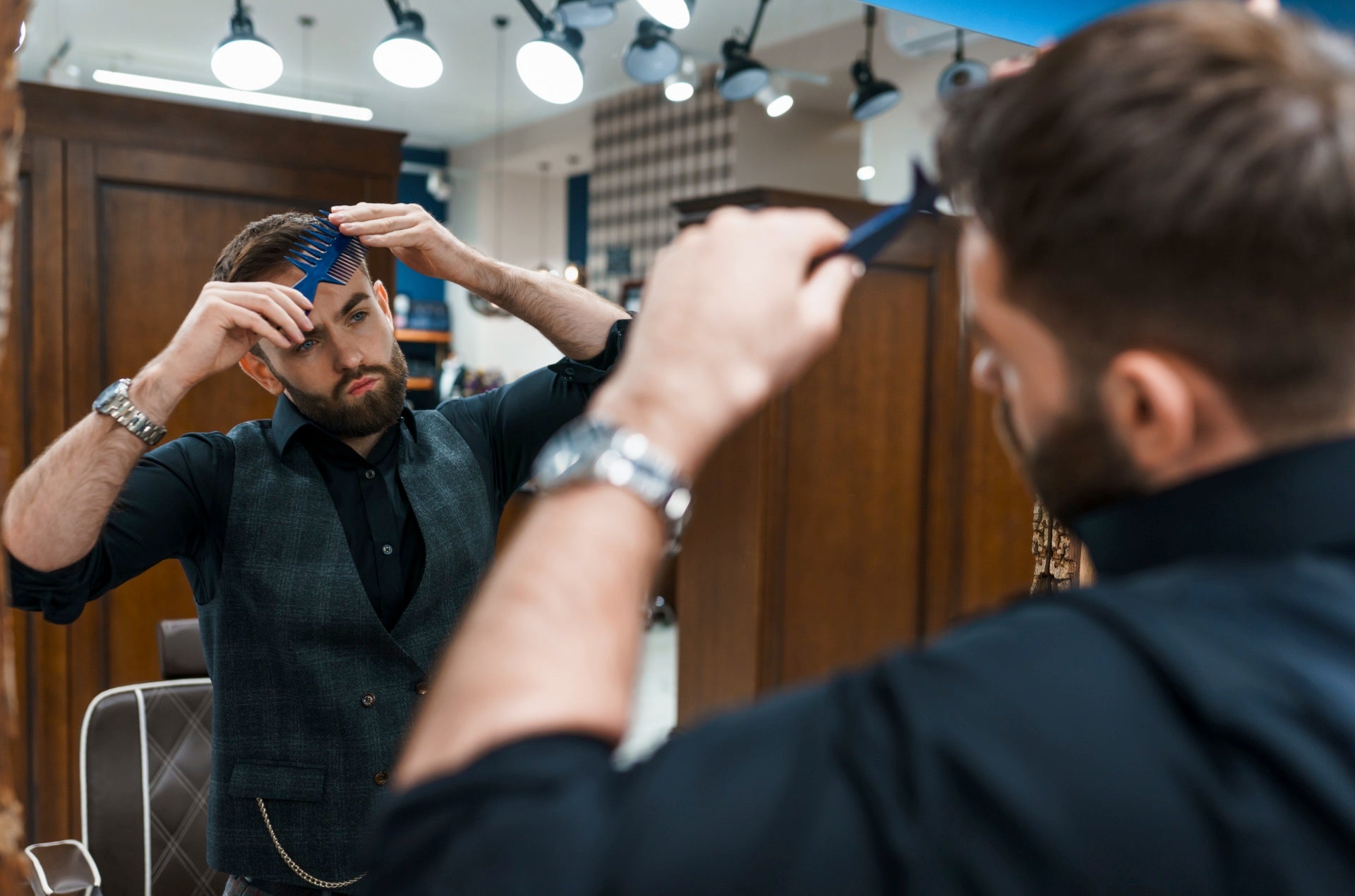 A man carefully adjusting his hairstyle using a blue comb, while checking his reflection in the mirror of a modern barbershop.