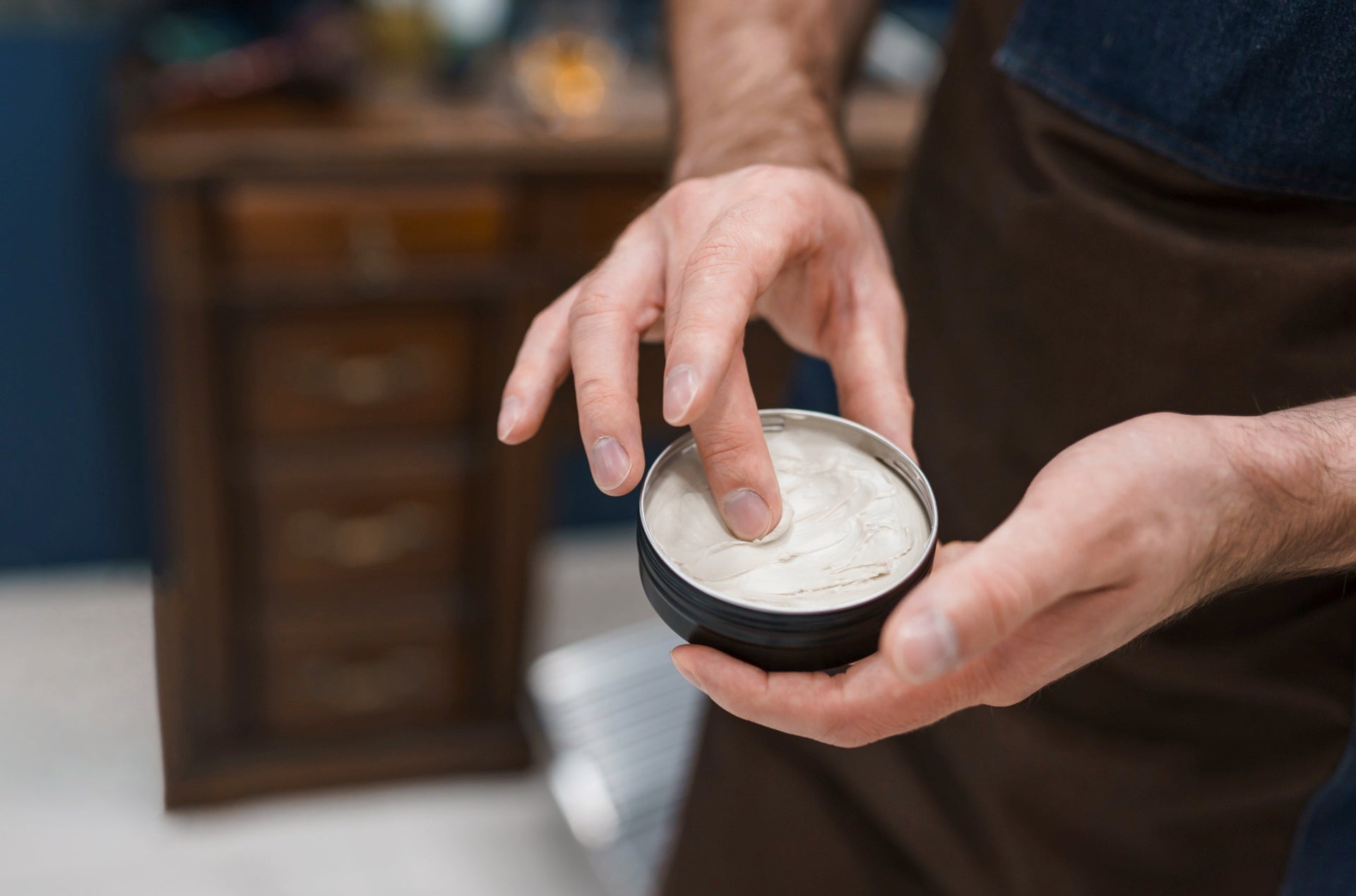 A man's hand dipping a finger into a tin of hair pomade, preparing to style his hair."