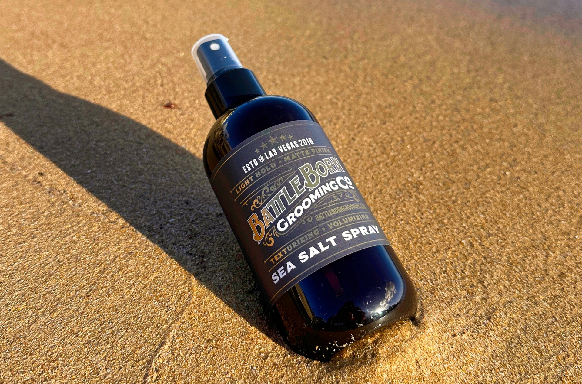 A bottle of Battle Born Grooming Co Sea Salt Spray nestled in a bed of sandy beach, evoking a sense of coastal freshness and natural beauty.