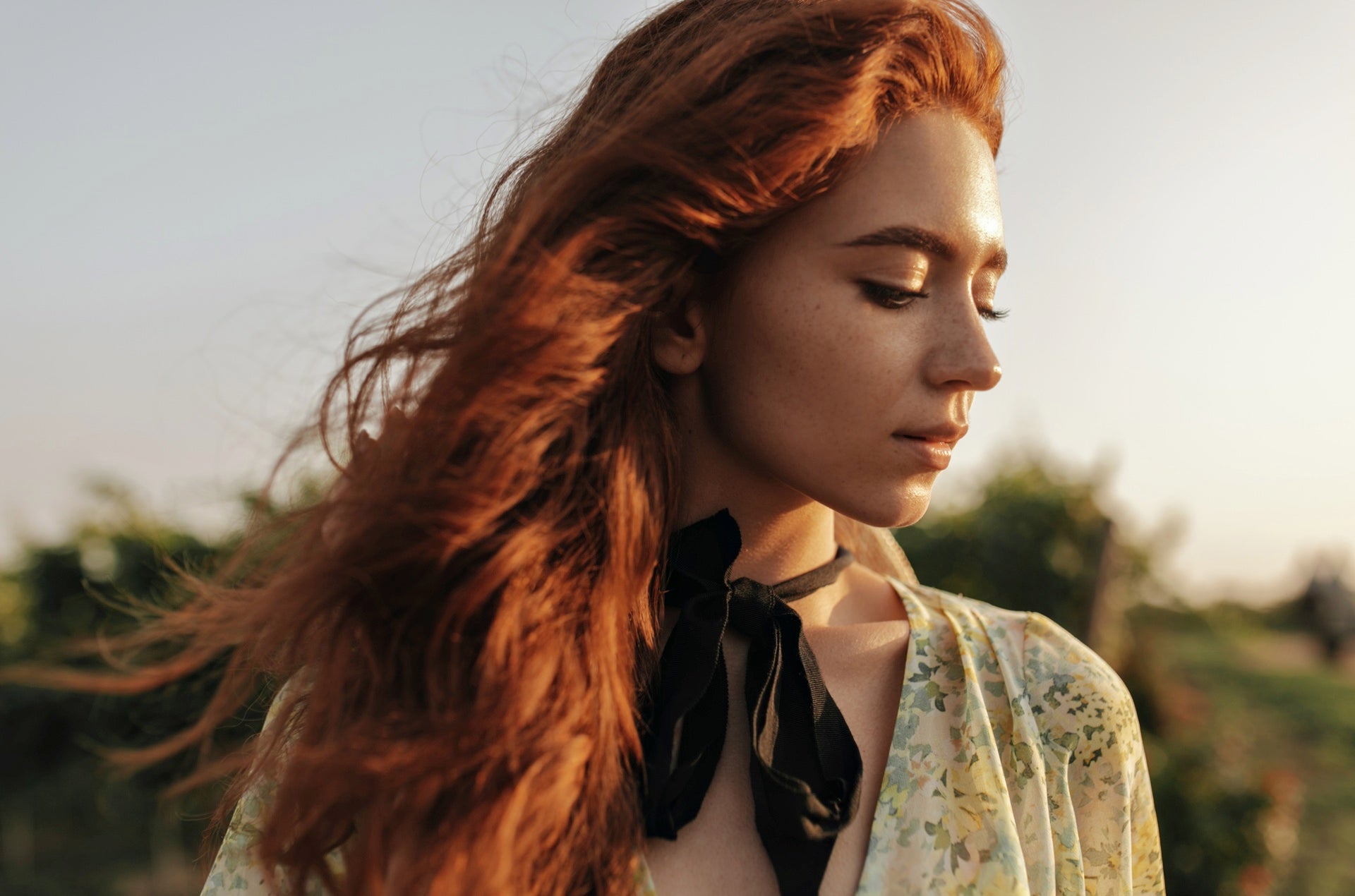 A woman with vibrant, wavy red hair bathed in golden sunlight. The gentle waves in her hair suggest the use of hair styling products to enhance its natural texture. A black ribbon is tied around her neck, and she wears a floral blouse, adding to the ethereal ambience of the image.