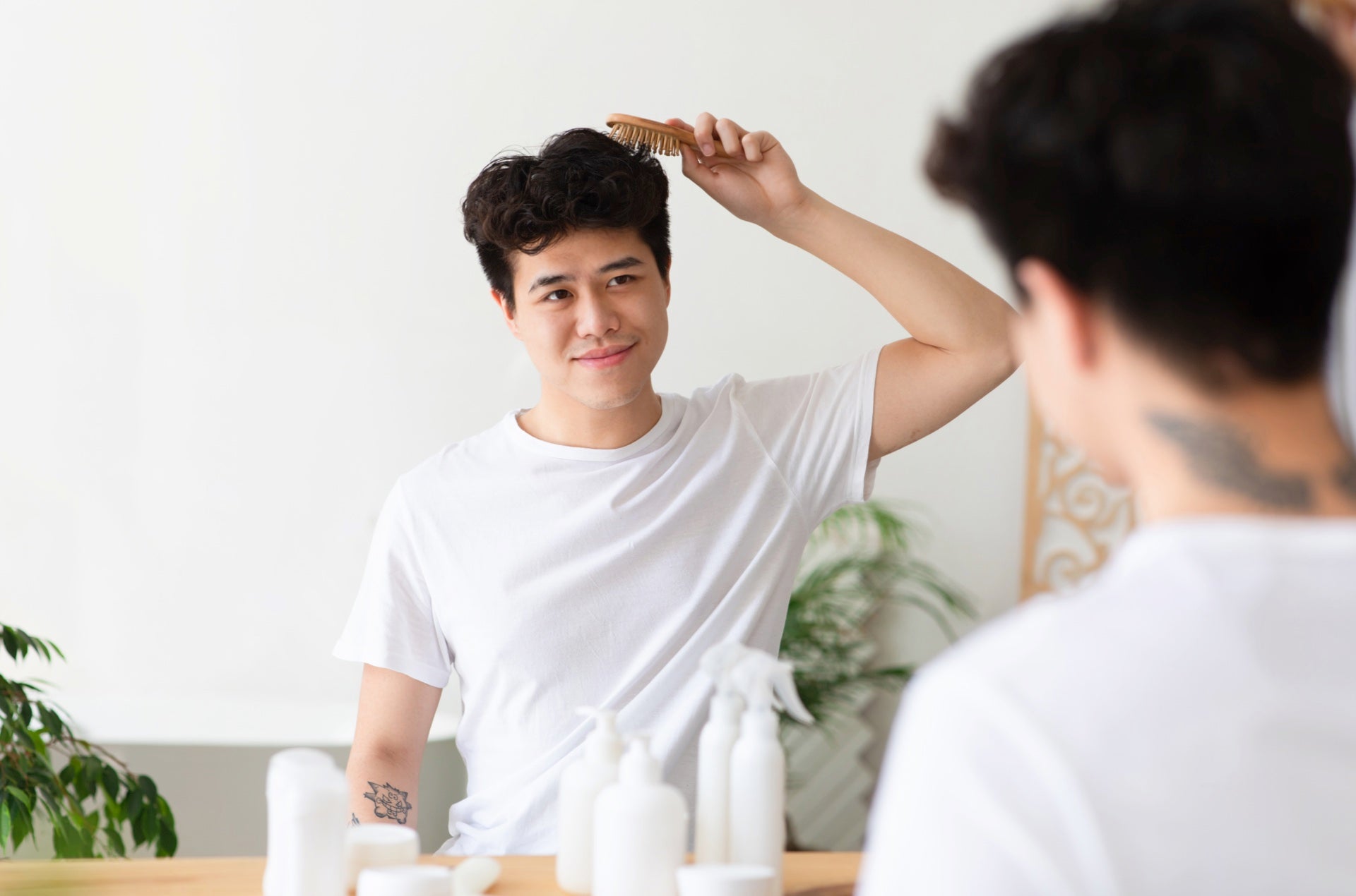 A young man with thick, curly hair uses a wooden comb to style it. Various hair styling products are neatly arranged on a table beside him, indicating his routine for maintaining and grooming his hair.