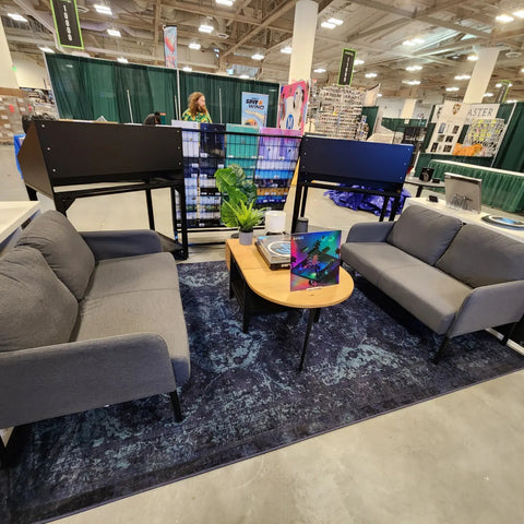 Materia Store booth at Emerald City Comic Con 2023. This image shows the lounging area of the booth with two couches and a table between them, sitting on top of a blue rug.