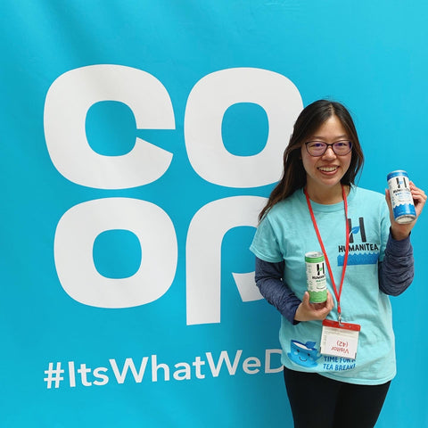 tina chen woman with Asian features and long dark hair and glasses wearing a turquoise teeshirt with a lanyard around her neck an black trousers and black long sleeved top undernath holding up two cans of tea standing in front of a COOP sign in turquoise with white lettering. Tina is the founder of HumaniTea.