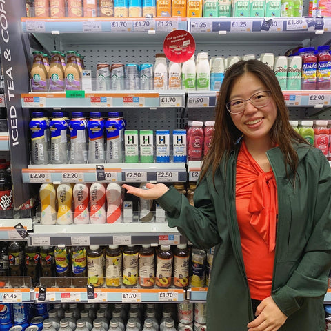 Tina Chen woman with Asian features and glasses smiling, wearing a red shirt with a khaki jacket holding up one hand in front of supermarket shelves featuring her brand HumaniTea
