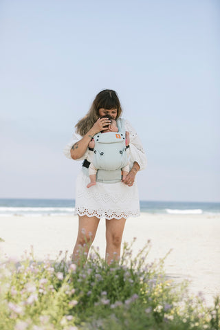 A mother who is carrying her child in a Tula Carrier and looking down at them while at the beach.