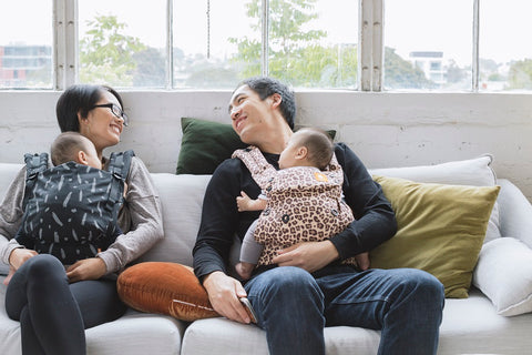 A babywearing couple sitting on a sofa with their children.