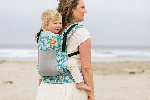 A mother carrying her child in a Coast Carrier in back-carry position, while standing in the shade.